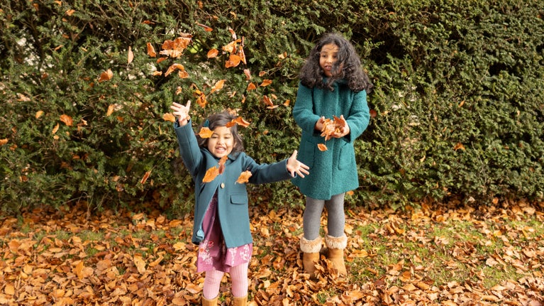 Two young girls in big coats are throwing autumn leaves in the air against the back drop of a yew hedge.